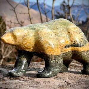 May include: A stone bear sculpture in shades of green, yellow, and black. The bear is depicted in a walking pose, with a smooth, polished finish. The sculpture is set against a blurred natural background.