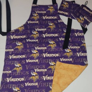May include: Purple apron and pot holders with a gold and white "Vikings" print. The apron has black straps and a gold-colored lining. The pot holders have a loop for hanging.