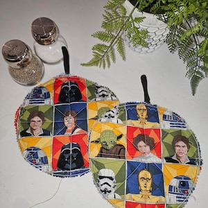 May include: Two round, quilted pot holders with a Star Wars theme. The pot holders are made of fabric with a patchwork design featuring characters from the Star Wars movies, including Darth Vader, Yoda, R2-D2, C-3PO, and Luke Skywalker. The pot holders are in a variety of colors, including red, blue, green, yellow, and black.
