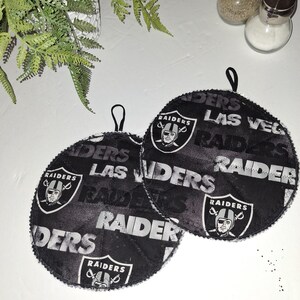 May include: Two black and white round pot holders with the Las Vegas Raiders logo and text. The pot holders have a quilted texture and a loop for hanging.