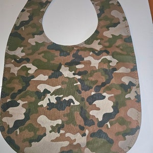May include: Camouflage fabric baby bib with a green, brown, and beige pattern. The bib has a rounded neckline and a curved bottom edge.