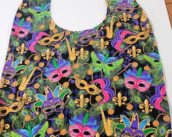 May include: A black bib with a Mardi Gras theme. The bib is covered in a repeating pattern of gold coins, green, purple, and gold feathers, and gold musical instruments. There are also multiple masks in various colors and designs.