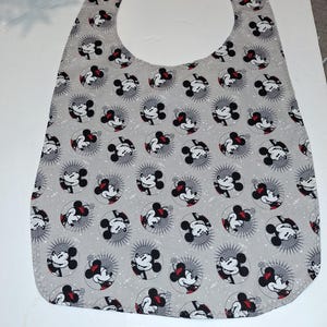 May include: A gray baby bib featuring a repeating pattern of Mickey and Minnie Mouse faces within circular designs. The bib has a rounded bottom and a wide neck opening, suitable for catching spills. The fabric has a light gray background with white and red accents.