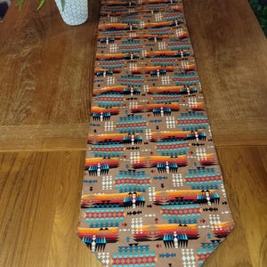 May include: A long, rectangular table runner featuring a repeating geometric pattern in shades of brown, orange, red, turquoise, and black. The fabric displays rows of stylized triangles, diamonds, and horizontal bands with pointed ends.