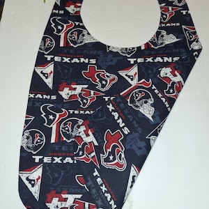 May include: A navy blue bib featuring a repeating pattern of the Houston Texans logo, helmets, and text. The bib has a rounded neck opening and a curved bottom edge. The team's logo is red, white, and blue.