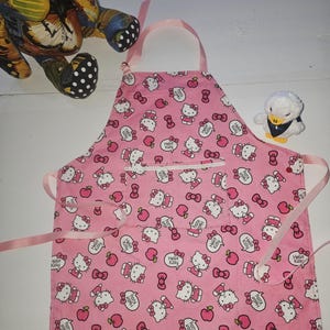 May include: Pink apron featuring a repeating pattern of Hello Kitty, bows, and apples. The apron has a pocket and pink ties. A plush duck and a decorative turtle are in the background.