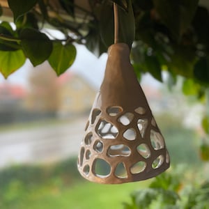 Könnte beinhalten: Eine weiße Keramik-Pendelleuchte mit geometrischem, durchbrochenem Design. Die Leuchte hängt an einem Ast mit grünen Blättern.