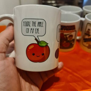 May include: White ceramic mug with a red apple illustration and the text "YOU'RE THE APPLE OF MY EYE." in a speech bubble.