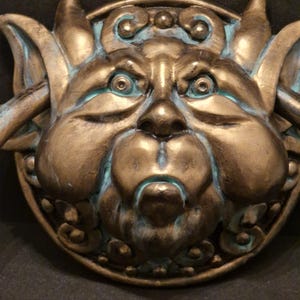 Puede incluir: Aldaba de puerta de bronce con diseño de gárgola y detalles turquesa. La aldaba presenta un rostro detallado con ojos grandes, mejillas prominentes y orejas puntiagudas. El diseño es circular con adornos decorativos.