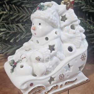 Puede incluir: Una figura de trineo de cer&aacute;mica blanca con un mu&ntilde;eco de nieve que lleva gorro y bufanda. El trineo est&aacute; decorado con formas de estrellas recortadas, detalles pintados en oro y peque&ntilde;as hojas de acebo. Una estrella dorada corona la estructura.