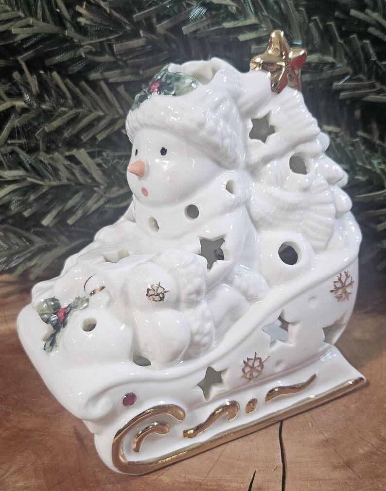 Puede incluir: Una figura de trineo de cer&aacute;mica blanca con un mu&ntilde;eco de nieve que lleva gorro y bufanda. El trineo est&aacute; decorado con formas de estrellas recortadas, detalles pintados en oro y peque&ntilde;as hojas de acebo. Una estrella dorada corona la estructura.