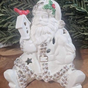Puede incluir: Una figura de Pap&aacute; Noel de cer&aacute;mica blanca con detalles de purpurina plateada en el gorro, la barba y el borde del traje. Pap&aacute; Noel sostiene una pajarera blanca con un p&aacute;jaro rojo y tiene un conejo al lado. El traje incluye una hebilla dorada.