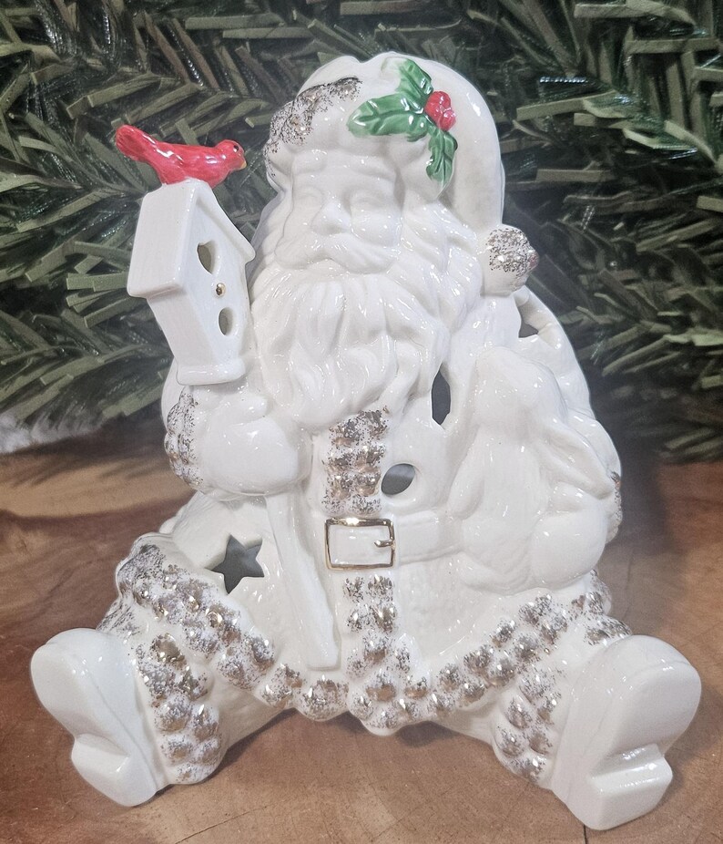 Puede incluir: Una figura de Pap&aacute; Noel de cer&aacute;mica blanca con detalles de purpurina plateada en el gorro, la barba y el borde del traje. Pap&aacute; Noel sostiene una pajarera blanca con un p&aacute;jaro rojo y tiene un conejo al lado. El traje incluye una hebilla dorada.
