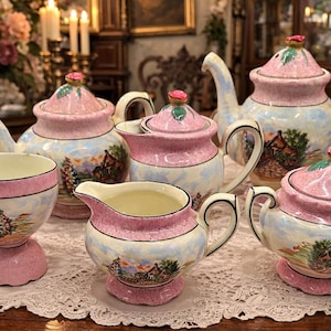 May include: A vintage tea set with a pink and white color scheme. The set includes a teapot, sugar bowl, creamer, and teacups. Each piece features a landscape design and is placed on a white lace doily.