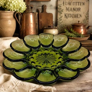 May include: A round, olive green glass serving platter featuring a scalloped edge with twelve individual recessed compartments surrounding a central starburst pattern. The pressed glass design has a smooth, reflective surface and a deep green hue.