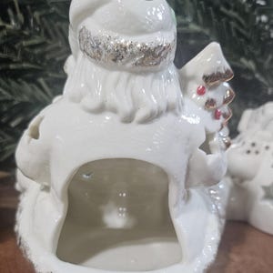 Puede incluir: Una figura de Pap&aacute; Noel de cer&aacute;mica blanca con la parte trasera hueca, con detalles de purpurina dorada en el gorro y la base, y un peque&ntilde;o &aacute;rbol con puntos rojos en la mano. La superficie brillante refleja la luz, resaltando la barba y el gorro.