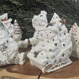 Puede incluir: Una colecci&oacute;n de figuras navide&ntilde;as de cer&aacute;mica blanca que incluye a Santa Claus, mu&ntilde;ecos de nieve y un trineo. Cada pieza presenta detalles en dorado y plateado, hojas de acebo y aberturas en forma de estrella para dejar pasar la luz.