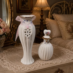 May include: A white ceramic vase with vertical fluting, a gold rim, and a pink ribbon tied around the neck with a heart-shaped cutout ornament, alongside a smaller white ceramic bottle with a heart-shaped stopper featuring floral patterns.