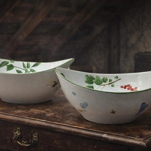 May include: Two white ceramic bowls with a unique, elongated shape and handles. Each bowl is decorated with green and red botanical illustrations and a green rim. The bowls are displayed on a dark wooden surface.