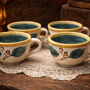 May include: Four ceramic coffee cups with white handles and a cream-colored base. Each cup has a teal interior, a yellow rim, and a painted leaf design. The cups sit on a lace doily on a wooden surface.