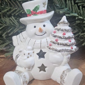 Puede incluir: Una figura de mu&ntilde;eco de nieve de cer&aacute;mica blanca que lleva un sombrero de copa con acebo, una bufanda y sostiene un regalo y un &aacute;rbol de Navidad. El mu&ntilde;eco de nieve presenta recortes en forma de estrella en el sombrero y el torso.