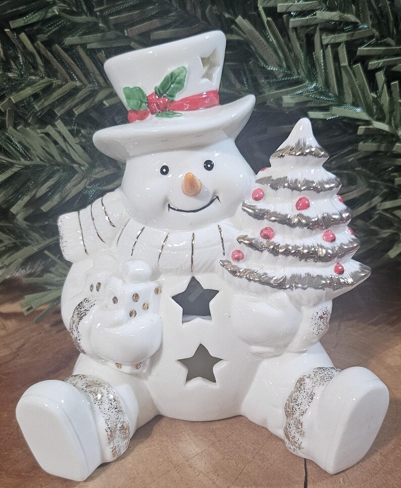 Puede incluir: Una figura de mu&ntilde;eco de nieve de cer&aacute;mica blanca que lleva un sombrero de copa con acebo, una bufanda y sostiene un regalo y un &aacute;rbol de Navidad. El mu&ntilde;eco de nieve presenta recortes en forma de estrella en el sombrero y el torso.