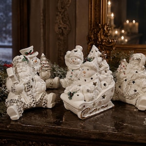 Puede incluir: Una colecci&oacute;n de figuras navide&ntilde;as de cer&aacute;mica blanca que incluye dos figuras de Pap&aacute; Noel, un mu&ntilde;eco de nieve en un trineo y un peque&ntilde;o mu&ntilde;eco de nieve con un &aacute;rbol. Cada pieza tiene detalles met&aacute;licos plateados, recortes de estrellas y acebo.