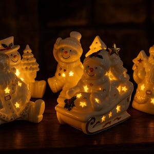 Puede incluir: Un conjunto de cuatro figuras navide&ntilde;as de cer&aacute;mica blanca, incluyendo a Santa Claus y mu&ntilde;ecos de nieve, con recortes en forma de estrella que emiten un c&aacute;lido resplandor amarillo gracias a una iluminaci&oacute;n interna. Las figuras est&aacute;n dispuestas juntas.