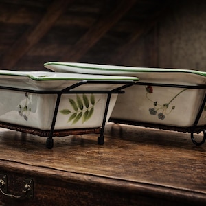 May include: Two rectangular ceramic planters with floral and leaf designs, set in black metal frames with decorative scrollwork. The planters have a white base with green trim and sit on a wooden surface.