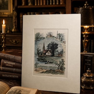May include: A rectangular off-white mat board framing a vintage-style color print. The print depicts a rural landscape with a church, a large tree, a house on a hill, and a river scene. The artwork is centered within the mat, which has a smooth, textured surface and clean, straight edges.
