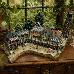 May include: A miniature seaside village diorama with buildings, a pier, beach, and a lighthouse. The buildings have colorful awnings and are surrounded by small trees. Miniature figures are on the beach and pier.