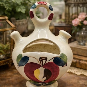 May include: Cream-colored ceramic decorative vase with a large opening and a handle with colorful polka dots. The vase features a painted apple design in red, yellow, and green, with a small base and two side spouts. A unique piece for home decor.