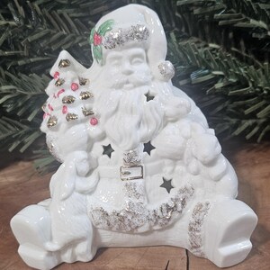 Puede incluir: Una figura de Pap&aacute; Noel de cer&aacute;mica blanca con detalles de purpurina plateada en el gorro, el cintur&oacute;n y las botas. Pap&aacute; Noel sostiene un peque&ntilde;o &aacute;rbol de Navidad con detalles dorados y rojos, y la figura incluye recortes en forma de estrella.