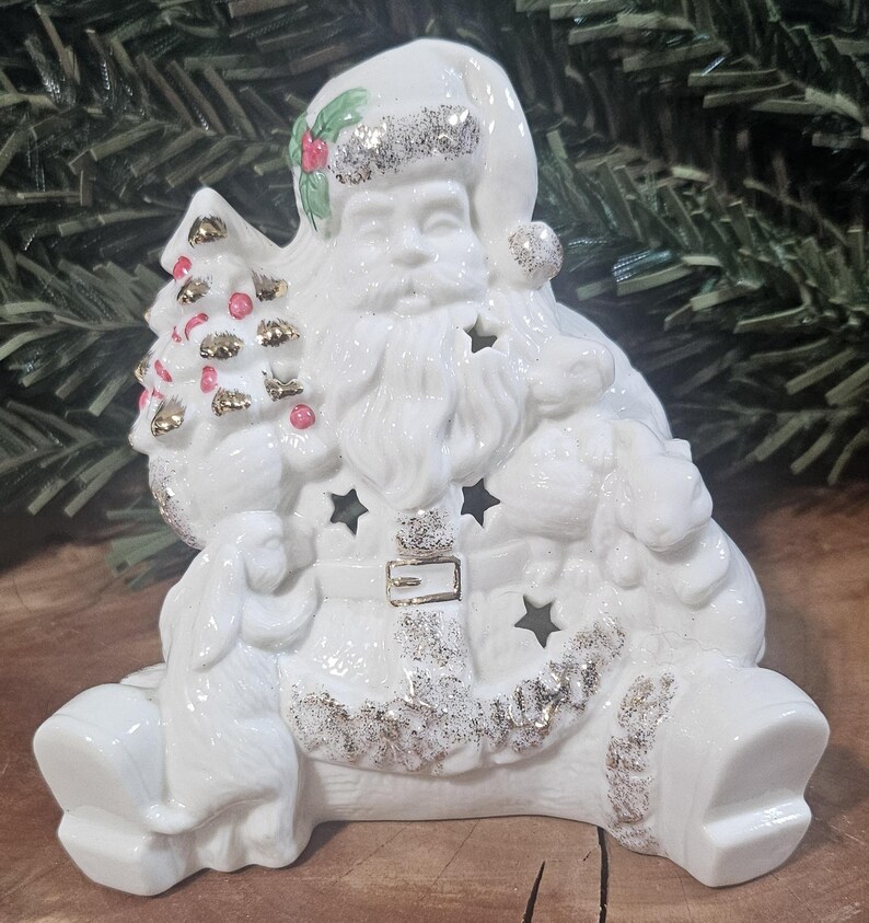 Puede incluir: Una figura de Pap&aacute; Noel de cer&aacute;mica blanca con detalles de purpurina plateada en el gorro, el cintur&oacute;n y las botas. Pap&aacute; Noel sostiene un peque&ntilde;o &aacute;rbol de Navidad con detalles dorados y rojos, y la figura incluye recortes en forma de estrella.