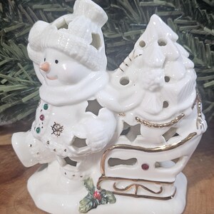 Puede incluir: Una figura de mu&ntilde;eco de nieve de cer&aacute;mica blanca con gorro y bufanda de punto, tirando de un trineo cargado con un &aacute;rbol de Navidad. La pieza presenta bordes dorados, recortes en forma de estrella y peque&ntilde;os detalles coloridos.