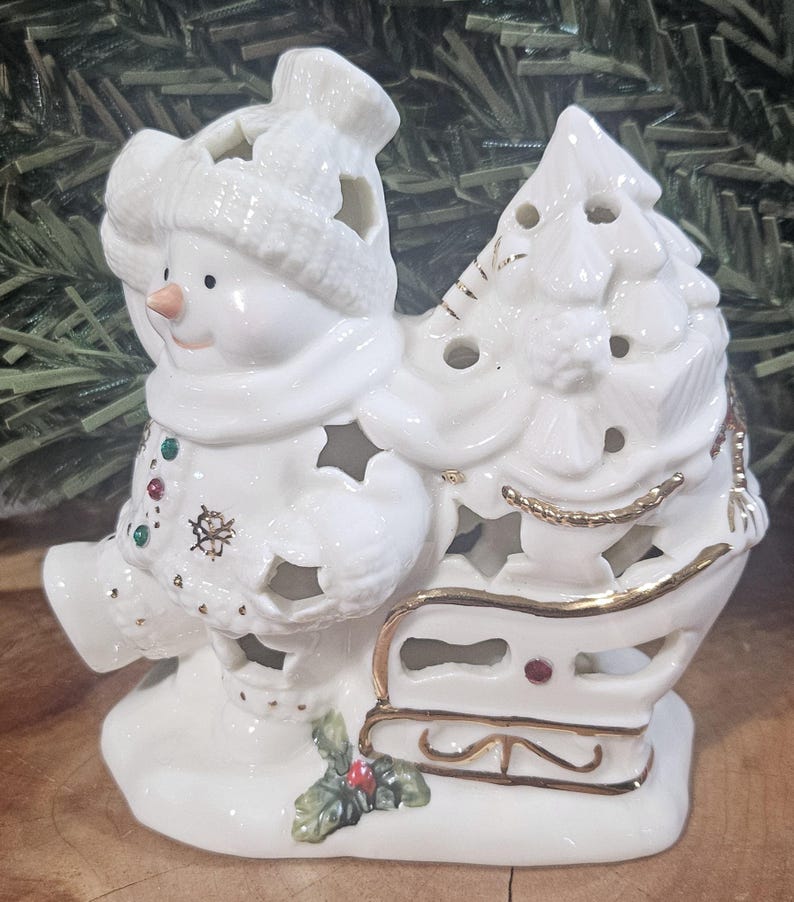 Puede incluir: Una figura de mu&ntilde;eco de nieve de cer&aacute;mica blanca con gorro y bufanda de punto, tirando de un trineo cargado con un &aacute;rbol de Navidad. La pieza presenta bordes dorados, recortes en forma de estrella y peque&ntilde;os detalles coloridos.
