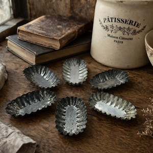 May include: A rustic still life featuring antique metal tart pans arranged on a wooden surface. The pans are oval-shaped with fluted edges. A vintage ceramic jar with "Pâtisserie" text and old books are also present.
