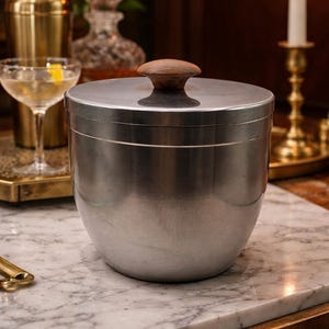 May include: A brushed metal ice bucket with a wooden handle on the lid. The ice bucket is cylindrical with a rounded base. A cocktail and a gold cocktail shaker are in the background. The ice bucket is on a marble surface.