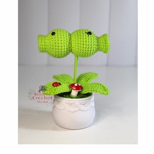 May include: A green crocheted plant with two round, green, crocheted flowers with black eyes. The plant is in a white pot with a lace trim and has a red mushroom and a red ladybug on the leaves.