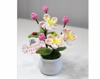 May include: A crocheted flower arrangement in a white pot with green leaves and pink and white flowers. The flowers have yellow centers. The arrangement is on a white surface. The text "Sesame Crochet World" is visible in the lower left corner of the image.