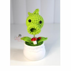 May include: A green crocheted plant with a large mouth and black eyes, sitting in a white pot with a lace trim. The plant has a red mushroom growing from the stem.