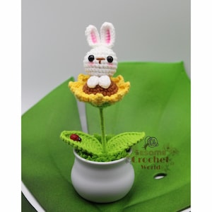 May include: A white crocheted bunny sitting in a yellow crocheted sunflower. The sunflower is in a white ceramic pot with green crocheted leaves. A red ladybug is on one of the leaves. The pot is sitting on a green surface. The text "Sesame Crochet World" is on the right side of the image.
