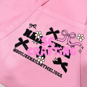 May include: A light pink apron featuring black embroidered text that reads NAILS BY MELISSA and @BOUJEENAILSBYMELISSA. The design includes black ribbon bow accents, white daisy flower icons, and a large pink ribbon graphic overlaying the centre.