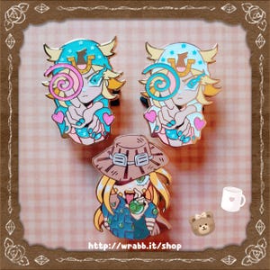 Puede incluir: Tres pines de esmalte con personajes de estilo anime. Dos pines muestran un personaje con un sombrero azul y un remolino rosa. El tercer pin presenta un personaje con un sombrero marrón. Los pines tienen contornos dorados.