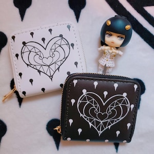 May include: Two square wallets, one white and one black, with a heart design. The heart is outlined in white and filled with geometric shapes. Small teardrop shapes are scattered around the heart. A small figurine stands behind the wallets.