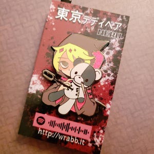 Może przedstawiać: Metalowa przypinka z postacią w stylu anime trzymającą czarno-białego misia. Przypinka ma japoński tekst na górze i adres URL http://wrabb.it na dole. Postać ma na sobie brązowy płaszcz.