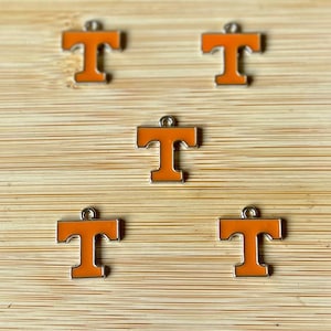 May include: Five orange letter "T" charms with silver-colored borders and small loops at the top. The charms are arranged on a light brown, wood-grain surface. The charms are a bright orange color.