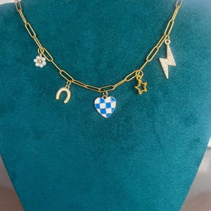 K&ouml;nnte beinhalten: Goldfarbene Kette mit mehreren Anh&auml;ngern. Zu den Anh&auml;ngern geh&ouml;ren eine Blume, ein Hufeisen, ein blau-wei&szlig; kariertes Herz, ein Stern und ein Blitz. Die Halskette wird auf einer teal-farbenen Samtoberfl&auml;che pr&auml;sentiert.