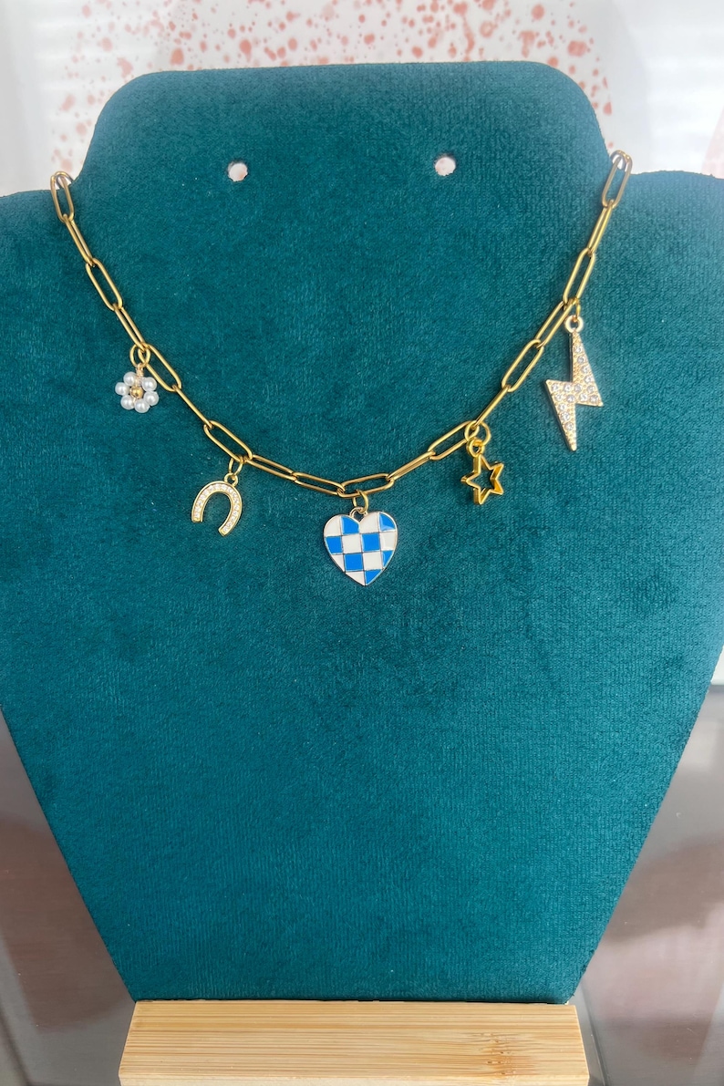 K&ouml;nnte beinhalten: Goldfarbene Kette mit mehreren Anh&auml;ngern. Zu den Anh&auml;ngern geh&ouml;ren eine Blume, ein Hufeisen, ein blau-wei&szlig; kariertes Herz, ein Stern und ein Blitz. Die Halskette wird auf einer teal-farbenen Samtoberfl&auml;che pr&auml;sentiert.