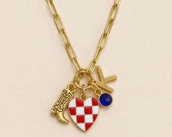 May include: A gold-tone paperclip chain necklace featuring a cluster of four charms: a gold cowboy boot, a heart-shaped charm with a red and white checkerboard pattern, a gold letter K, and a small circular charm with a deep blue gemstone.